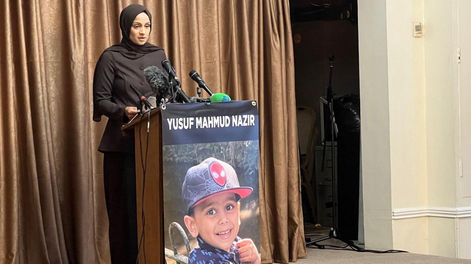 A woman with her hair covered stands in front of a lectern with the picture of a young boy smiling on it. It has the words Yusuf Mahmud Nazir at the top of the lectern above the image.