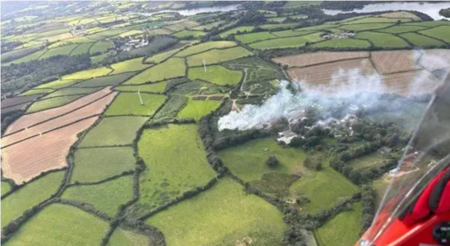 Aerial image of smoke belching upwards into the atmosphere from a compost fire on the ground which is surrounded by green and verdant arable crop fields and in the distance the gentle bend of a river or lake along with an enticing glimpse of a light plane fuselage painted in a rich and attractive red hue.