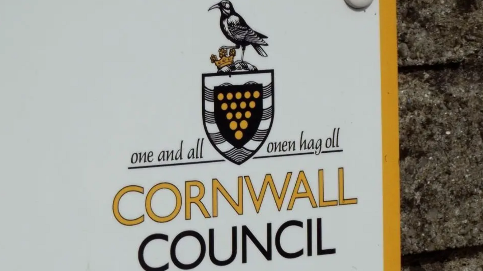 White and yellow sign reads "One and all Cornwall Council". The sign is on a brick wall.