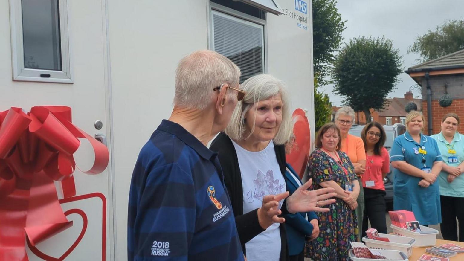 A white van is in the background with a large red ribbon around it. To the right, people are standing and watching, as a man and a woman stand in front of the bus. The woman has grey hair and is wearing a white T-shirt. The man is in a navy-striped top and has grey hair. He is wearing sunglasses.