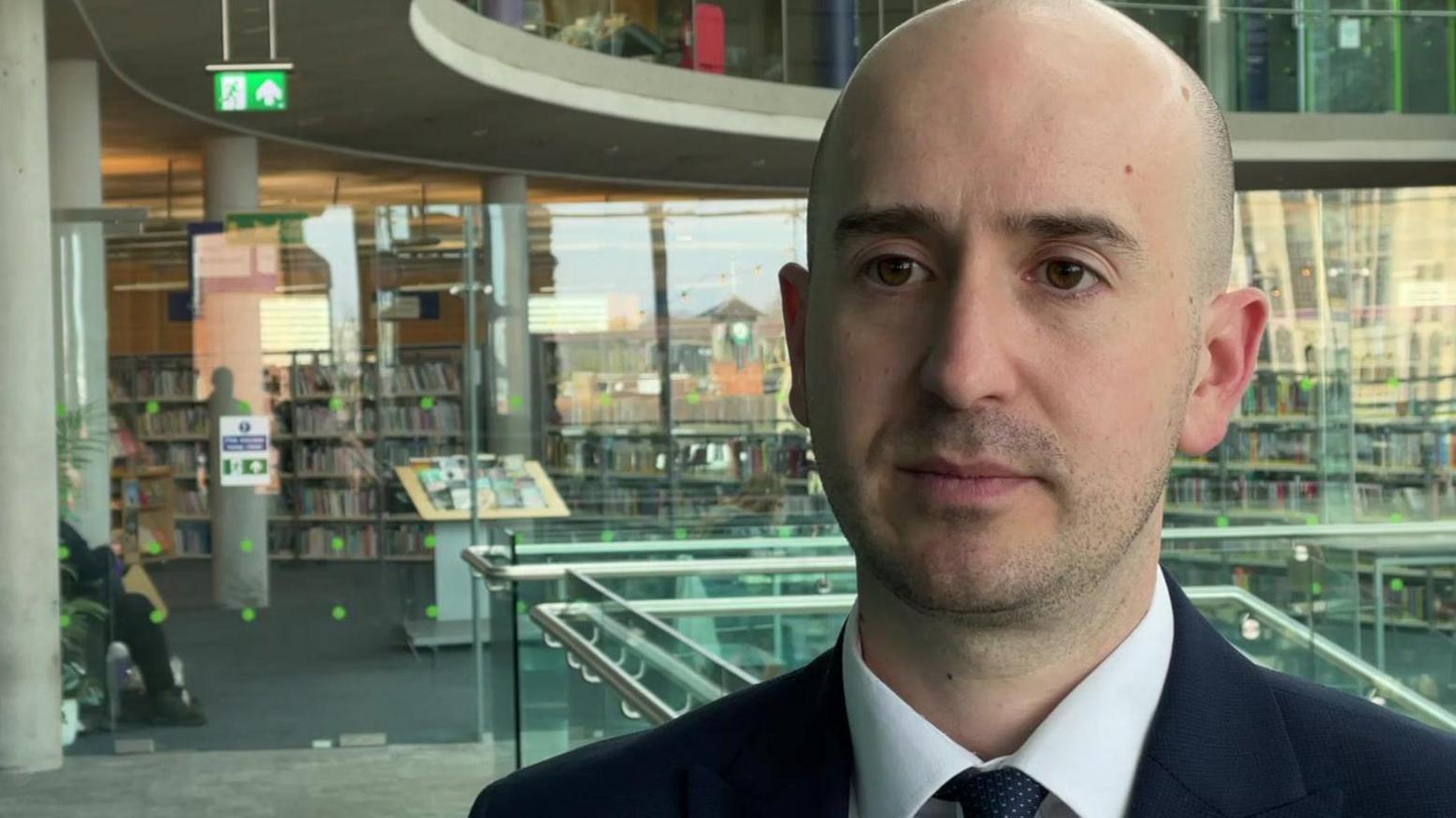 Ben Goldsborough is standing just off centre to the right, and is facing the camera. He is wearing a blue suit jacket, a matching tie and a white shirt. Behind him is a library behind a glass wall.