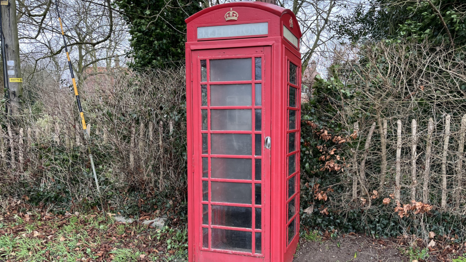 A traditional red phone box stands by a wooden fence with trees behind it.