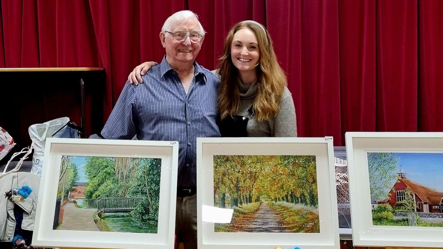 Robert Rose and granddaughter Joanna Menzies standing behind three white-framed prints of his paintings - a bridge, path through autumn leaves and a church