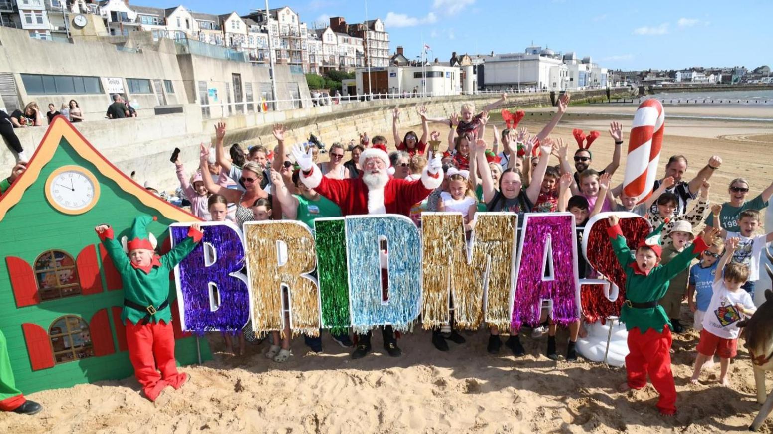 Beachgoers stand in the sand and cheer with a glittery 'Bridmas' sign, with elves and Father Christmas joining in the celebration.