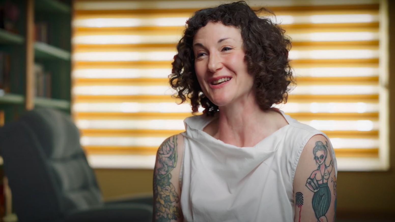 Imogen, wearing a white top, smiles. She has multiple tattoos on both of her upper arms.