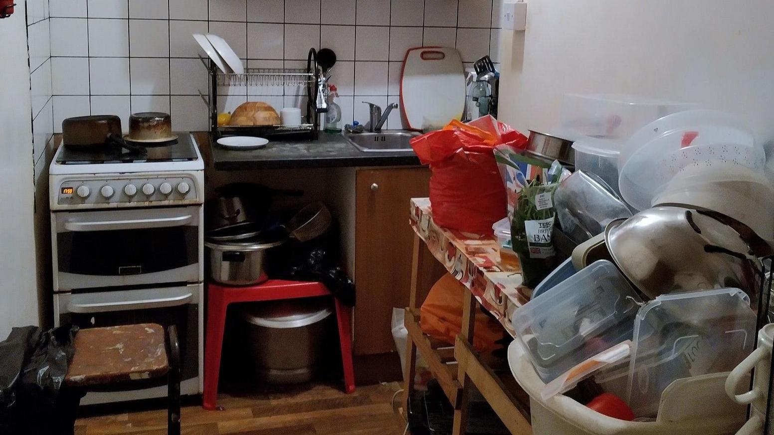 Image shoes a small kitchen overflowing with food and cooking equipment, including a large stack of tupperware, pans and bowls. There is only one small oven and sink, and a number of very full plastic bags in the room. 