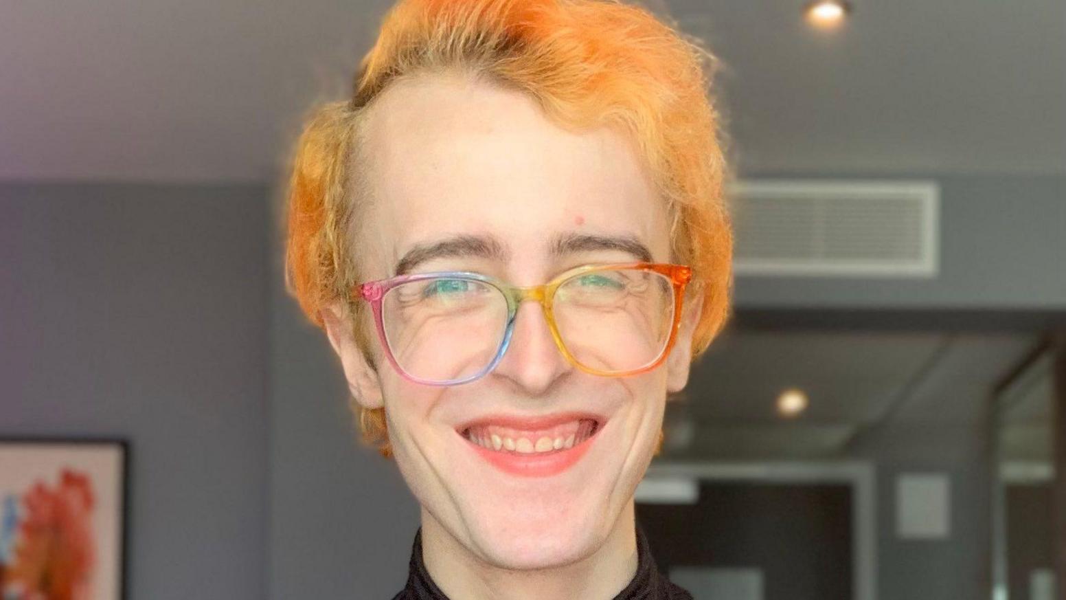 Casey Byrne has orange hair and rainbow glasses. He is smiling at the camera.