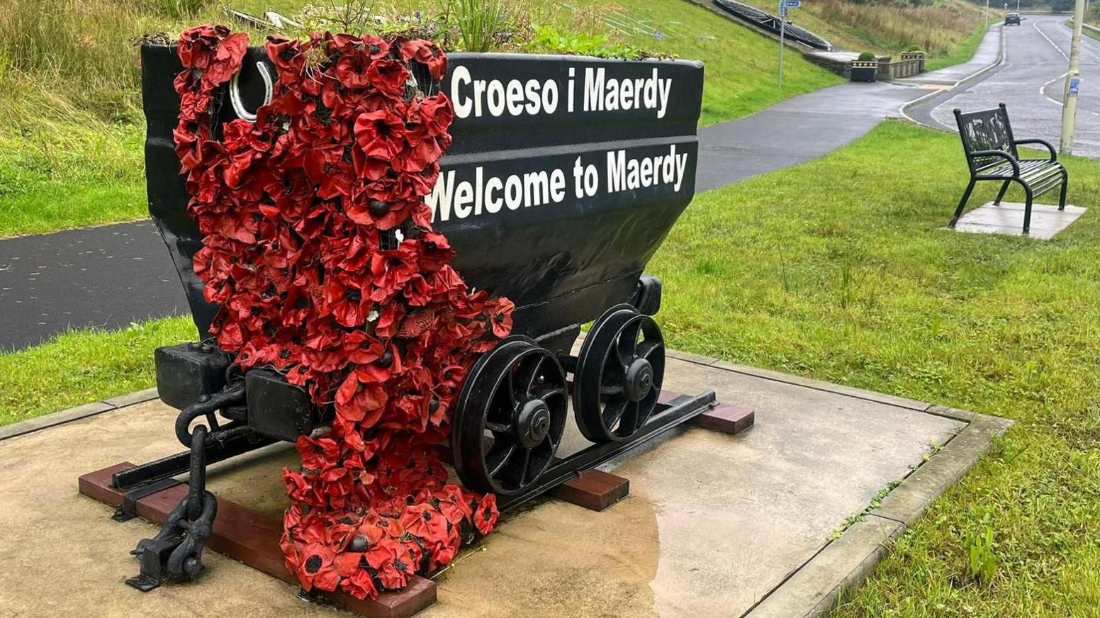 A metal mining cart which says 'welcome to Maerdy' and has fake poppies decoratively draped from it