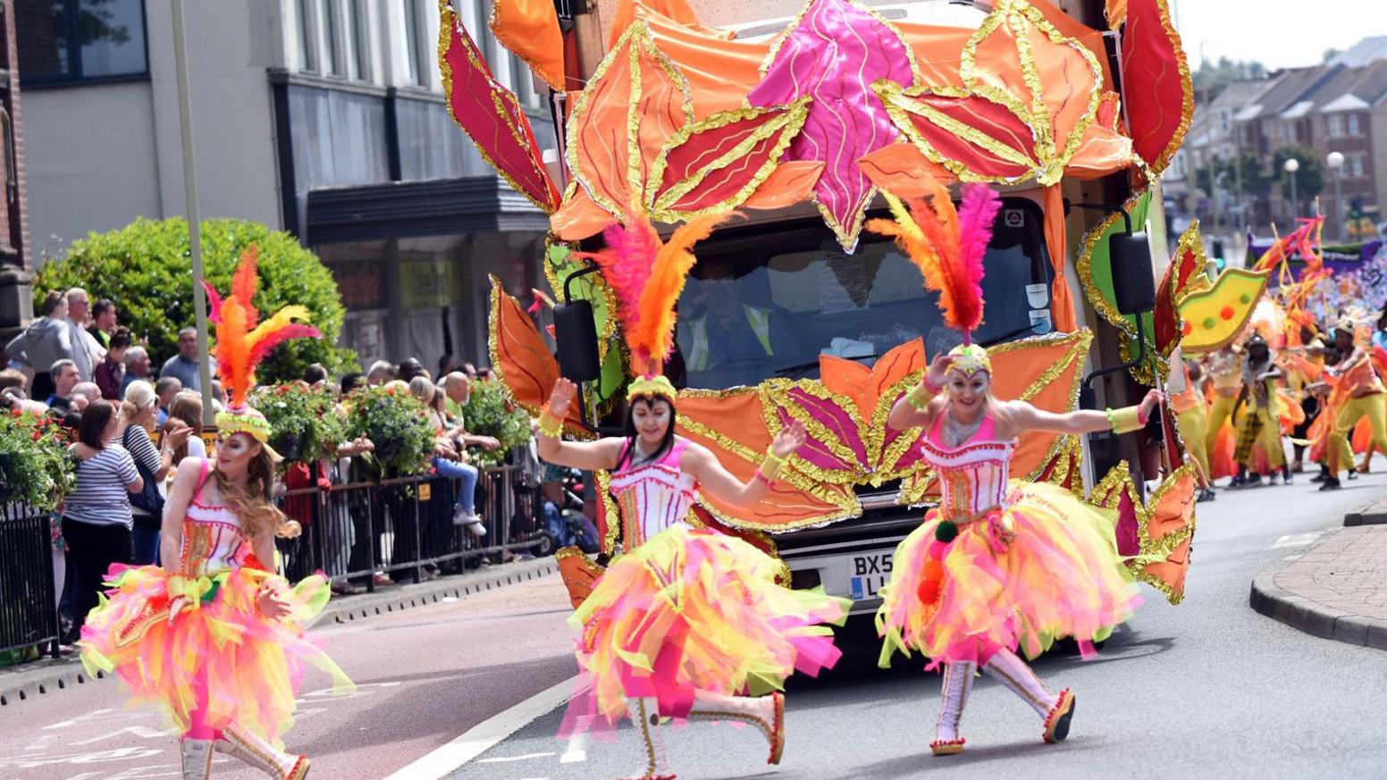 Three female dancers are performing in the middle of a road as part of a parade. They are wearing pink and yellow tutus and carnival clothing, including large feathers on their heads. A bus is behind them covered in orange, pink and gold leaves. The rest of the carnival procession can be seen behind them.