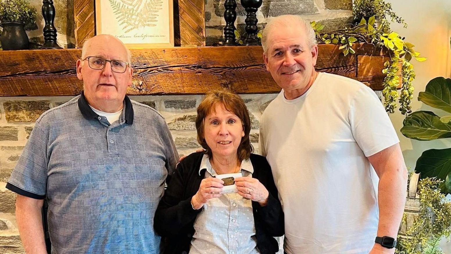 Bridgette Daily and her husband, next to Donald Madar on the right, wearing a white T-shirt. Bridgette in the middle is smiling holding up the dog tags. Her husband has a blue polo shirt on and wears glasses.