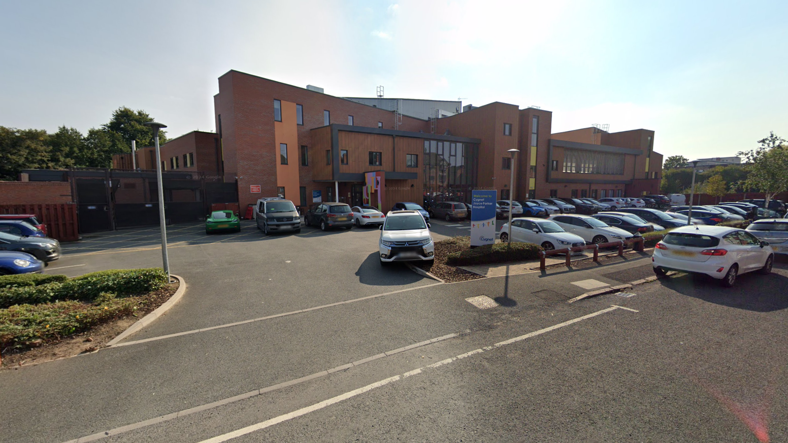 A Google Streetview image from the hospital, a modern brick and glass building,  with cars parked outside. 