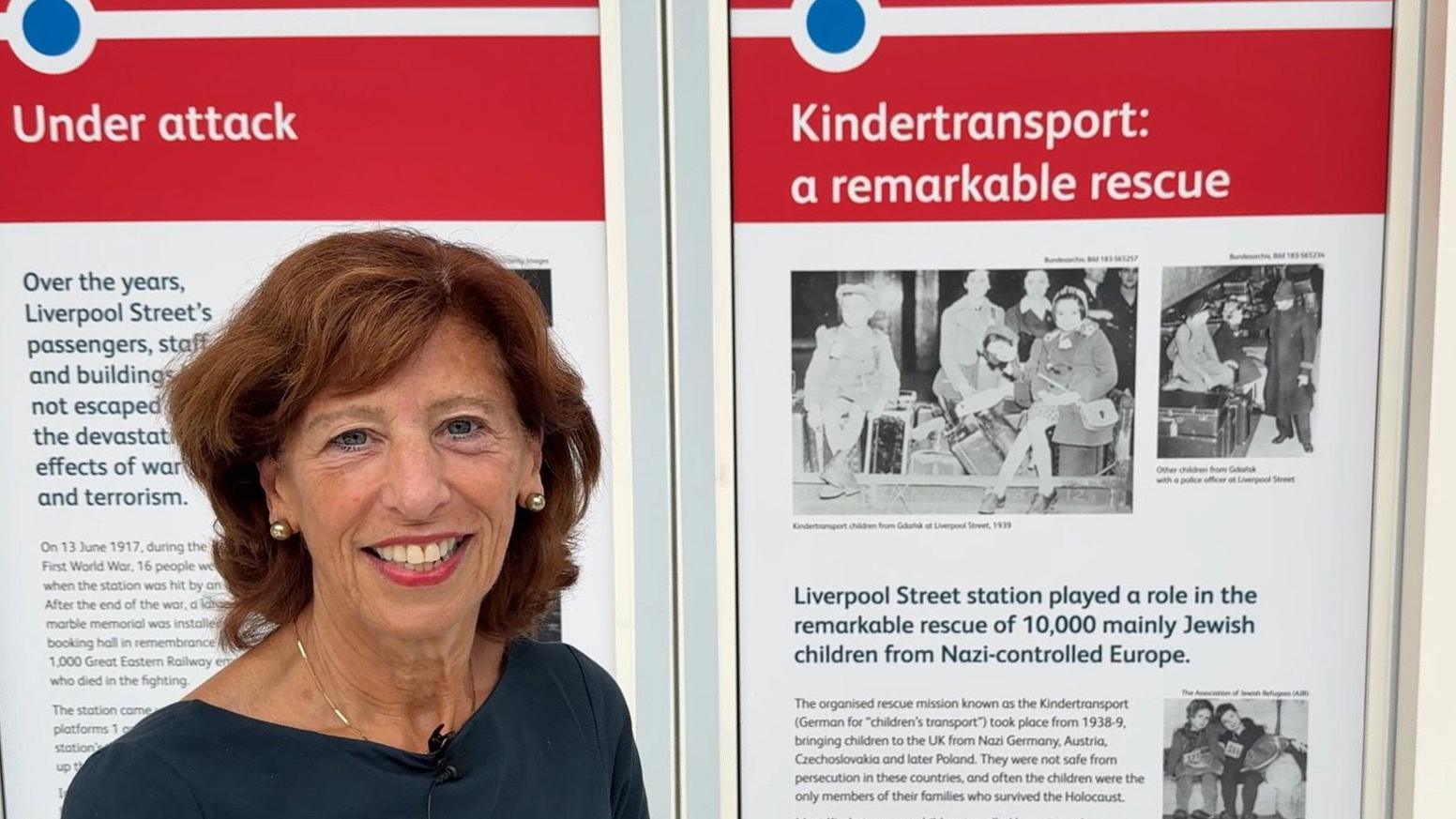 Lady Linda Reich in a dark coloured top standing in front of a display board about the history of the Kindertransport.