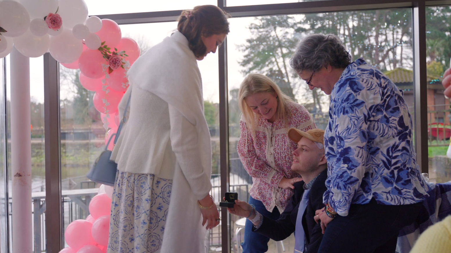 Charlie is on one knee proposing to Judy. His mums are supporting him. There are pink balloons in the background.