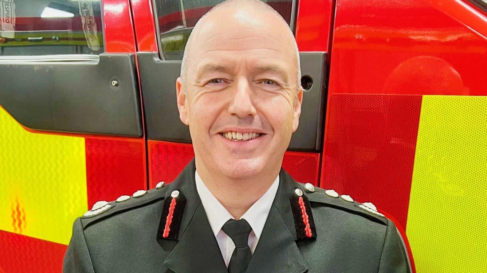 Mark, a bald man with a bright smile, wears his fire uniform as he stands in front of a red and yellow fire engine.