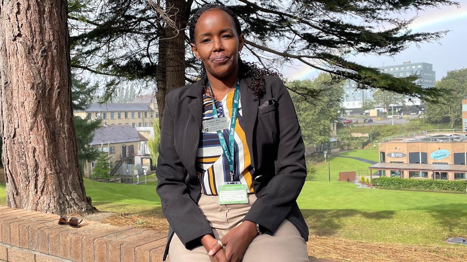 A woman sat on a brick wall with a university campus behind her. She wears a black jacket, beige trousers and a yellow, blue and shite patterned top.