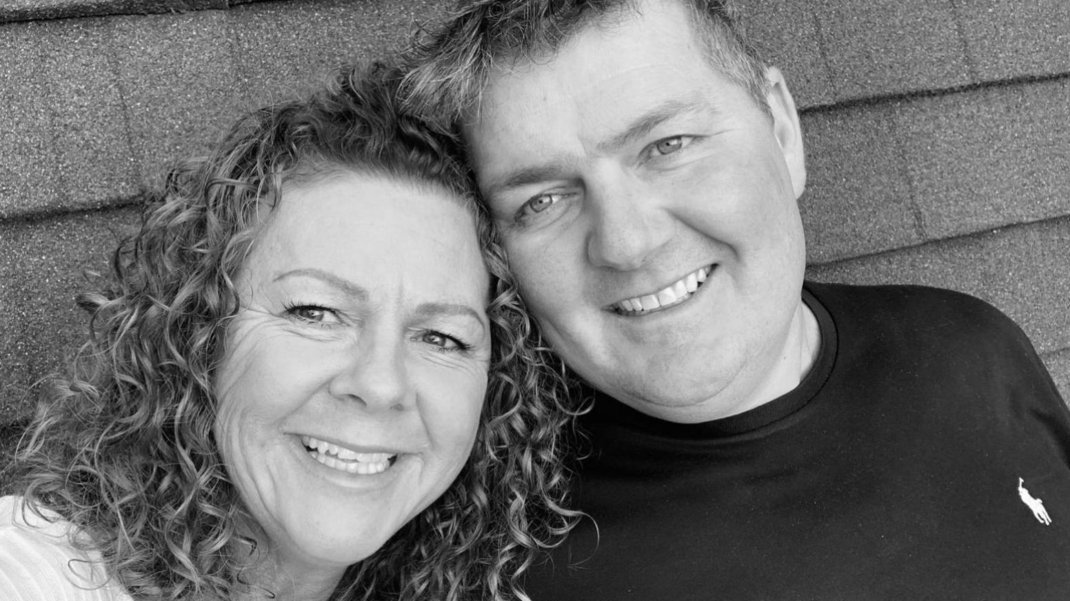 Jane Hutchinson on the left with curly hair and looking at the camera, smiling with her husband James. The photo is black and white. James is wearing a black Ralph Lauren t-shirt and is smiling fondly. He has blonde hair and is of stocky build.