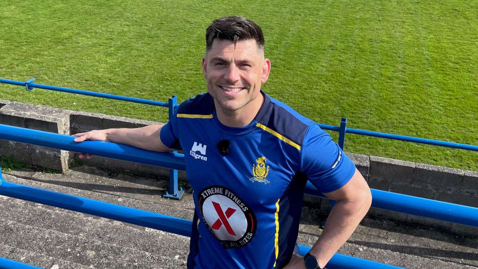 James Newton, smiling and holding on to a blue standing rail at Whitehaven's ground in a blue and yellow Whitehaven training top