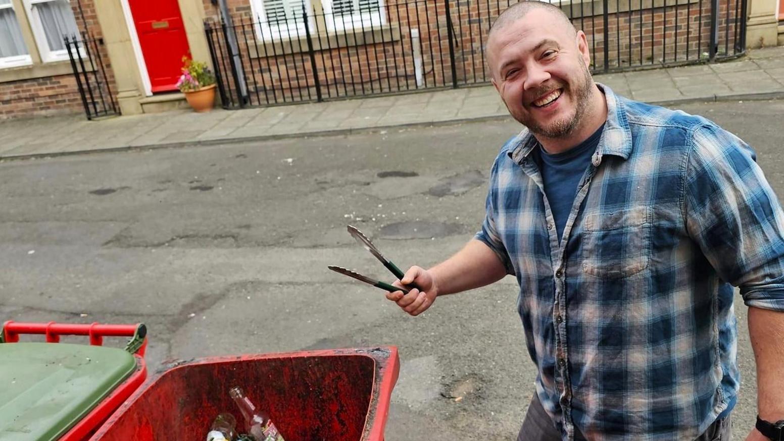 Smiling man holding a pair of tongs while standing in a street next to a red glass recycling bin