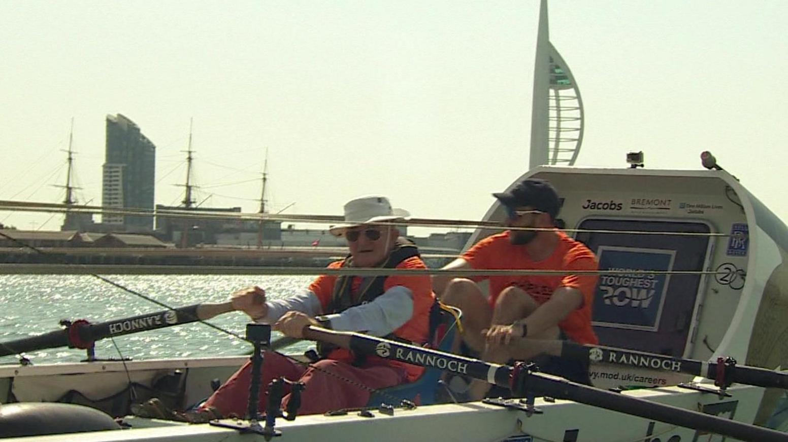 Two men sitting in a boat, using oars to row. In the background is the Spinnaker Tower.