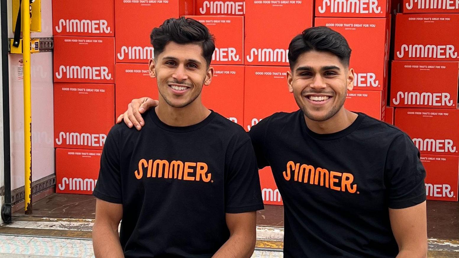 Two men of South Asian heritage men smile at the camera. They are each wearing black T-shirts with the word "Simmer" in orange lettering on them. Behind them is a stack of red food delivery boxes bearing the same logo in white.