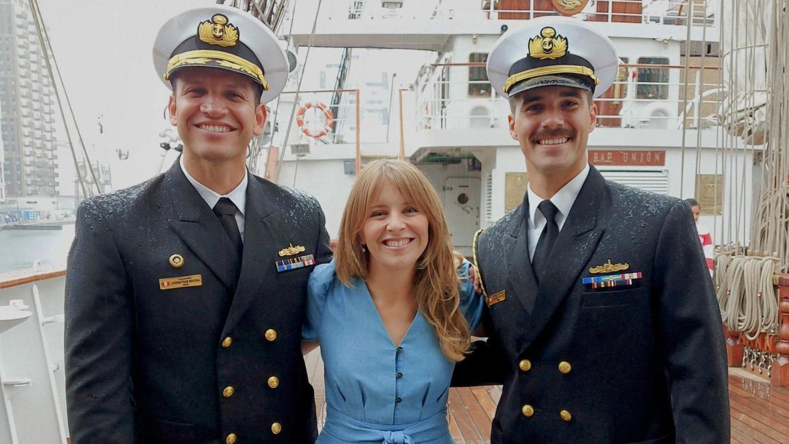 Two naval officers wearing smart uniforms and white military hats stand in the rain either side of a BBC reporter wearing a blue jumpsuit, on the wooden deck of a tallship, with ropes wound neatly behind them