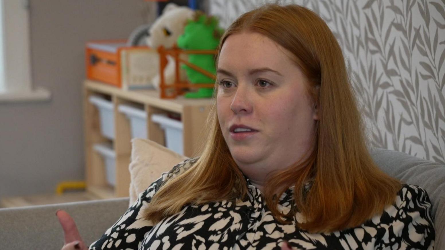 Alex Fearnley had long red hair and is wearing a black and white patterned top. She is sat on a grey sofa in her living room with children's toys in the background.