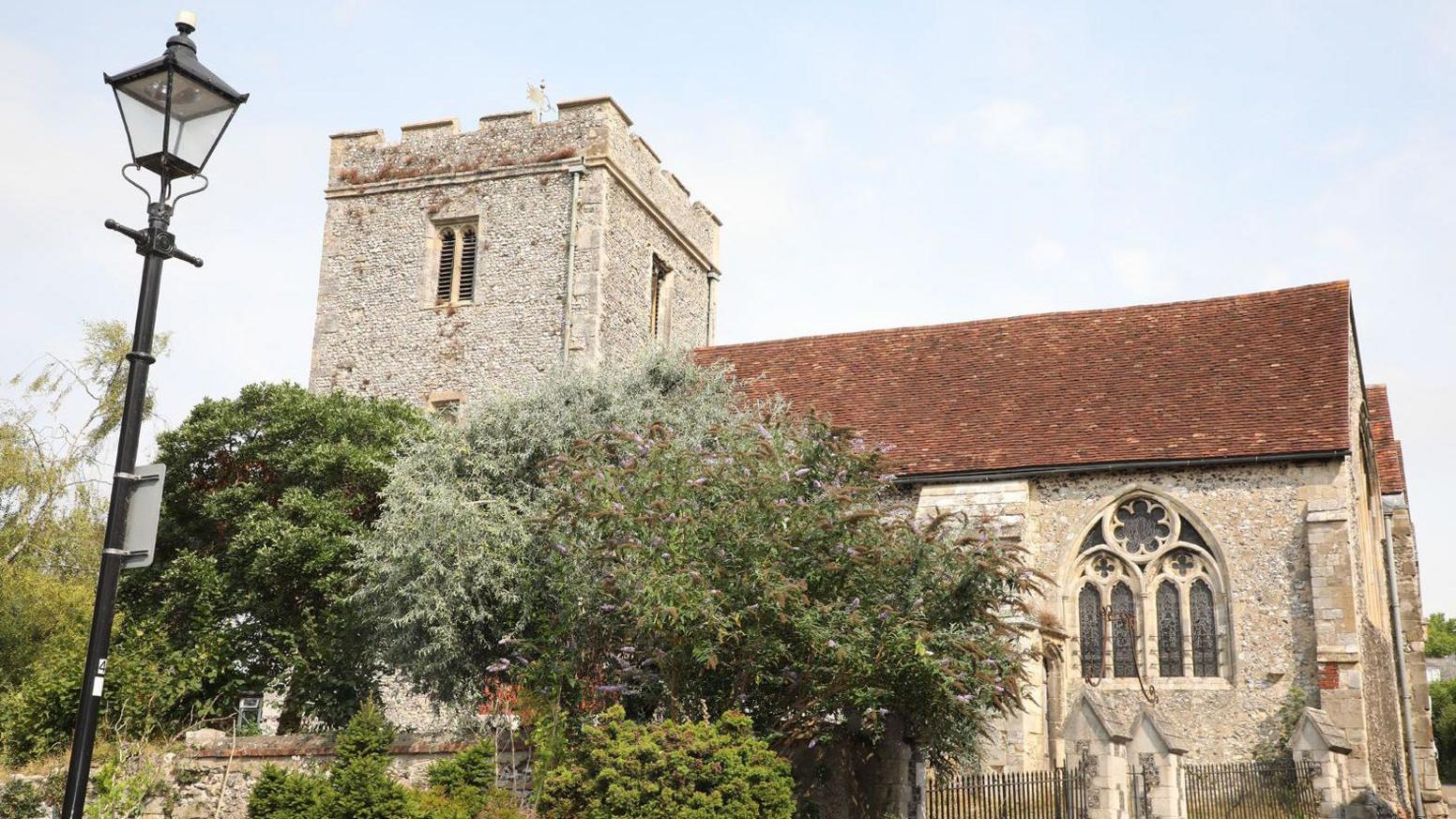 The church with its flint walls and red-tiled roof is bathed in sunshine with a gold weather vain on top of the bell tower.