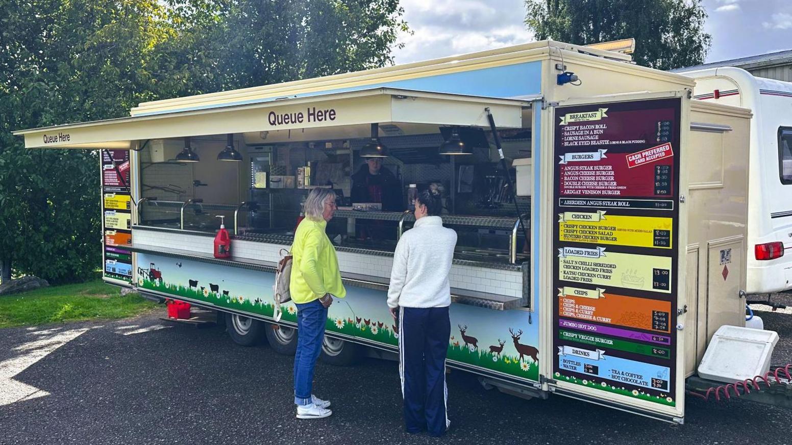 A food truck, with brightly coloured menus on it, with two women standing in front of it.