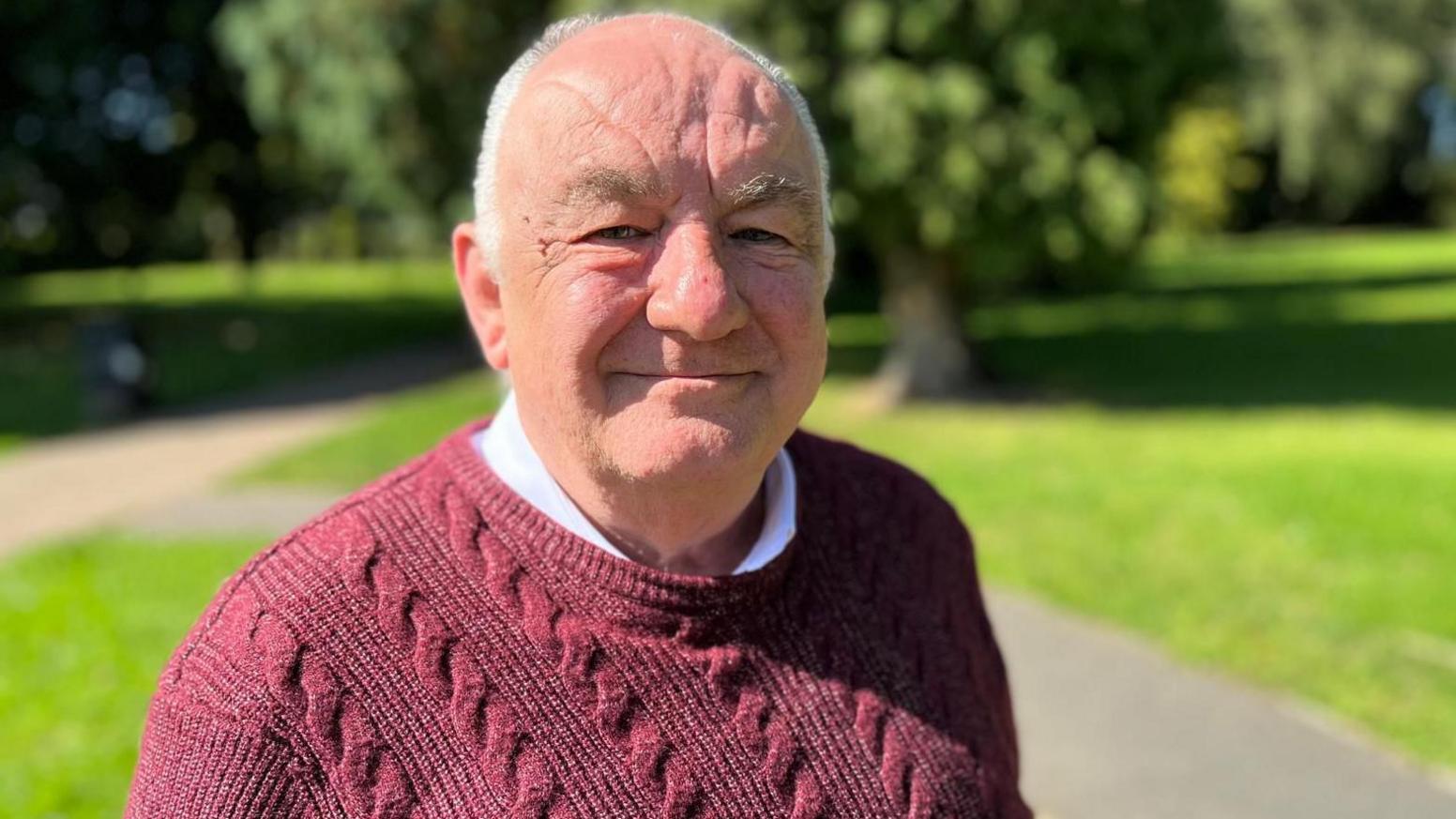 Alan Slade smiling in bright early autumn sunshine in a public park with leafy green trees behind him and wearing a red jumper