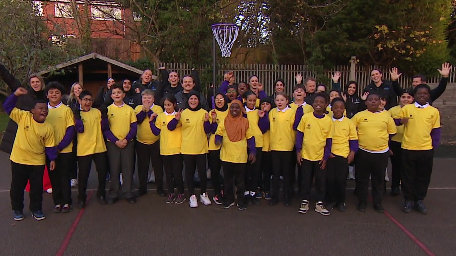 More than a dozen pupils stand in a line in yellow shirts on their playground waving and smiling at the camera. Netball players and school staff stand behind them. A netball net can be seen behind them all