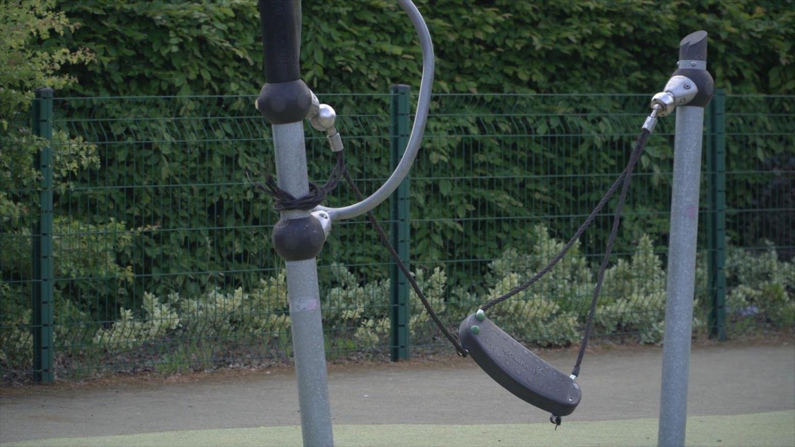 A children's swing in a playpark. One of the four ropes on the swing is broken.