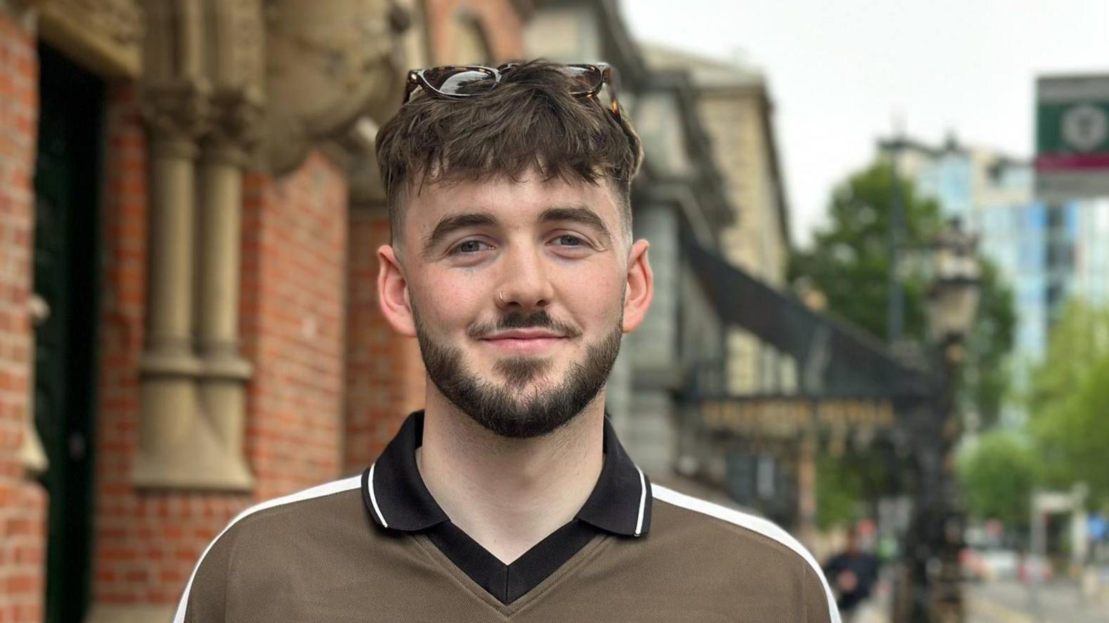 A young man with short brown hair, dark moustache and beard. He is wearing a green polo shirt. He has sunglasses on his head.