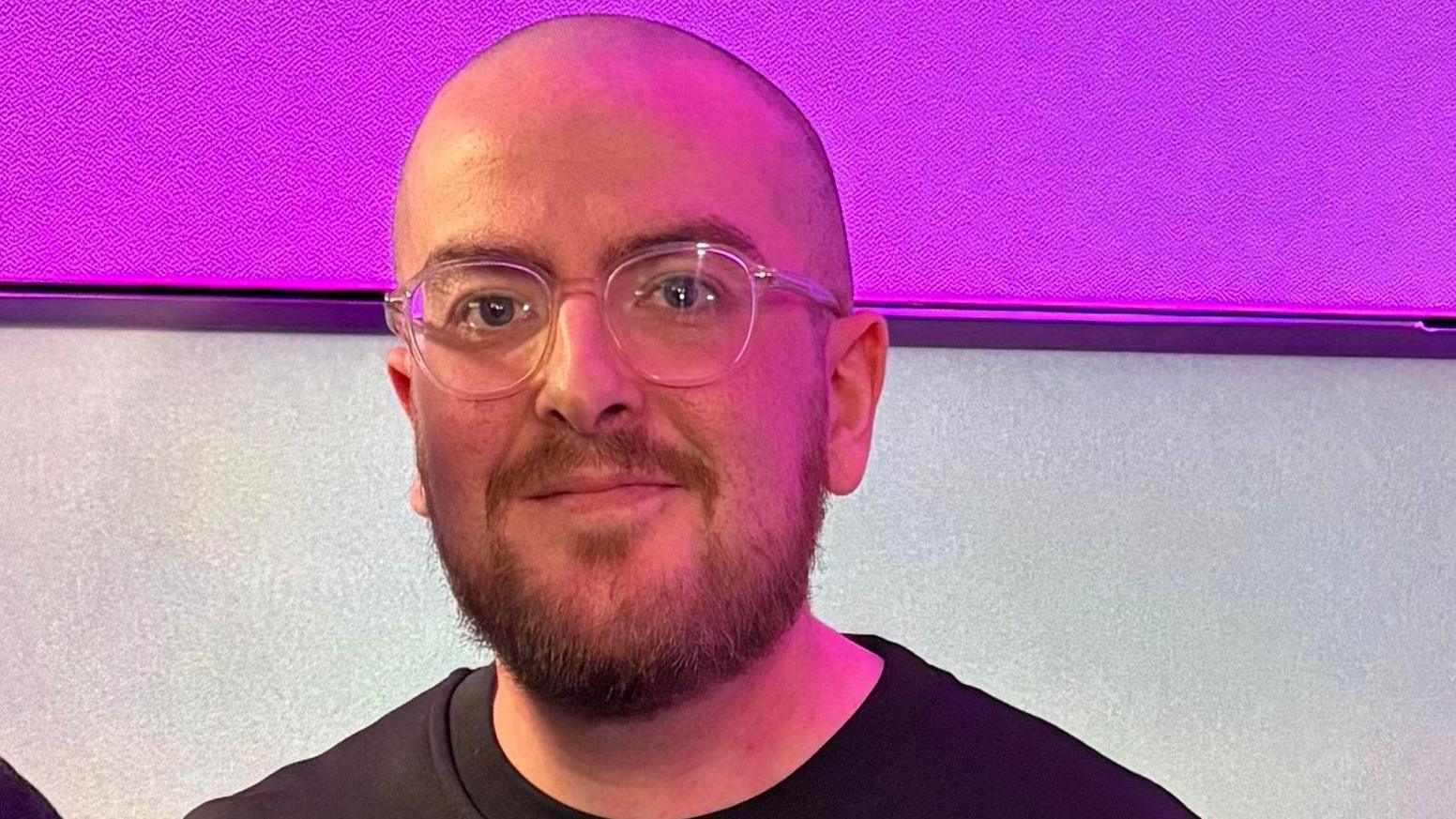 A man wearing glasses and a black jumper smiles as he stands in a studio with a purple and grey wall behind him.