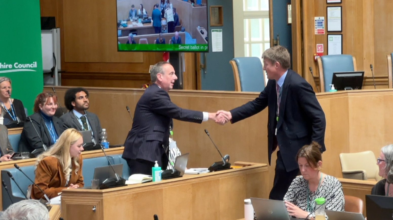 New leader Ian Thorn (left) shakes hands with former leader, Conservative Richard Clewer (right) while in the council chamber. They have other councillors sitting around them in a space that is a little like a lecture theatre with lots of wooden panelling