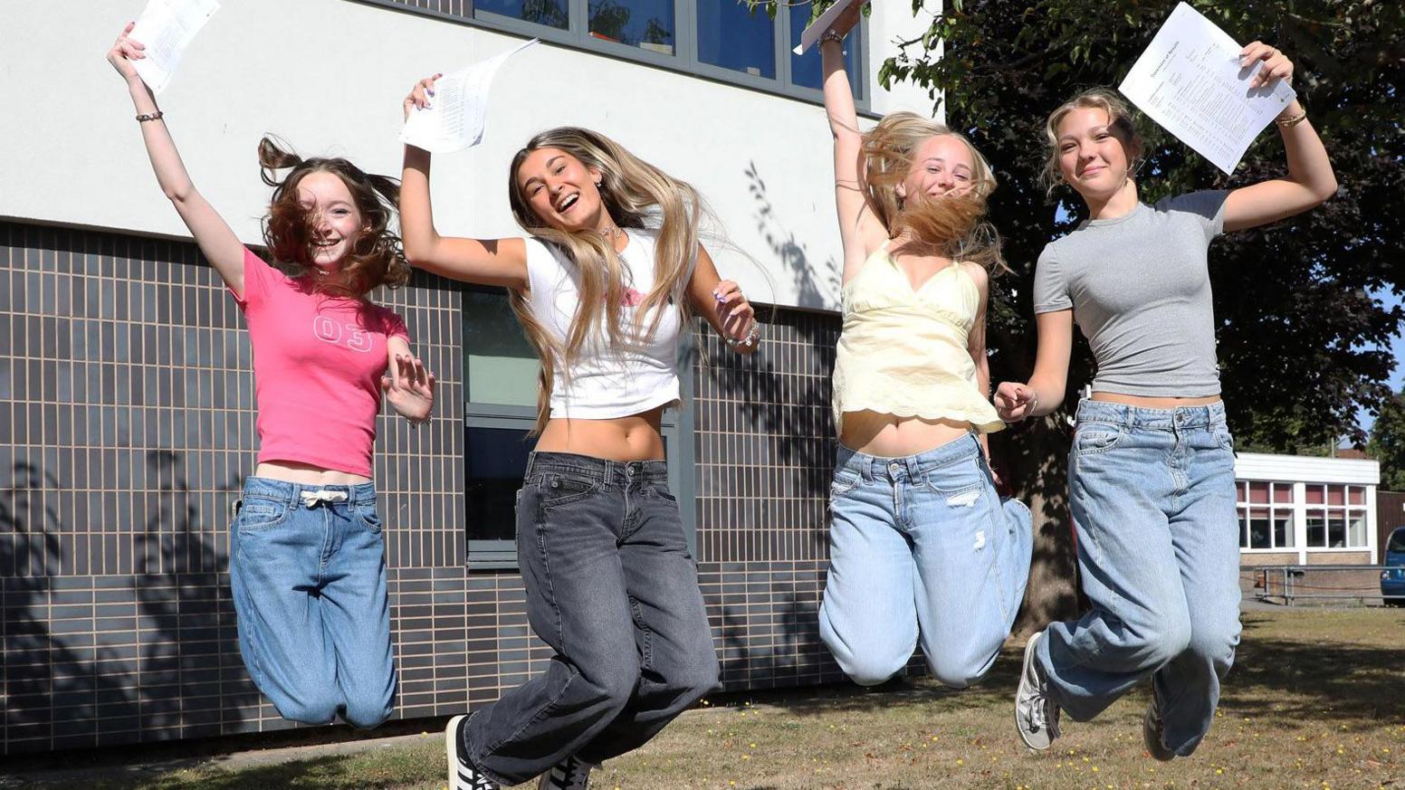 Four young women are jumping up in the air, holding a page with their exam results. They are all smiling, their knees are slightly bent as they are mid-leap. The girls, who are all wearing casual t shirts with baggy fit jeans, are outside of their school, there are school buildings behind them and some patches of grass.