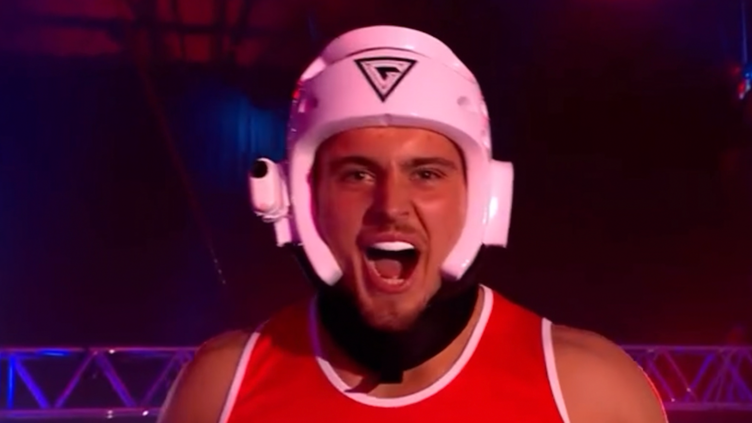 A contestant in Gladiators wearing a white helmet and a red shirt with the logo.