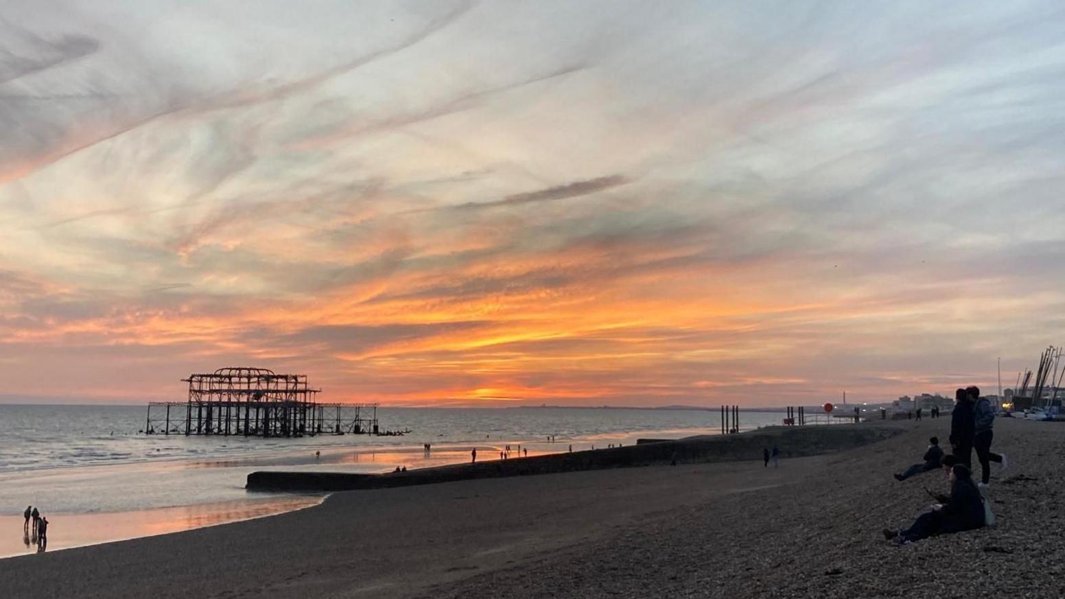 A picture of the sea and the beach at sunset with layers of high cloud in the sky, a few people sitting on the beach and the ruins of Brighton's burnt out West Pier visible.