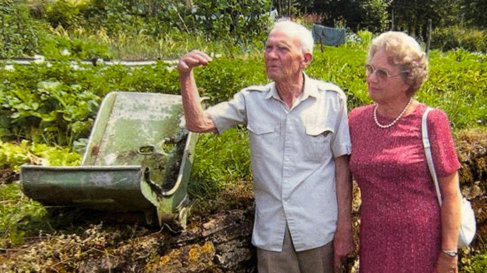 An older man and woman standing next to a grass verge. On the verge is a green piece of metal, which is the shell of a seat.