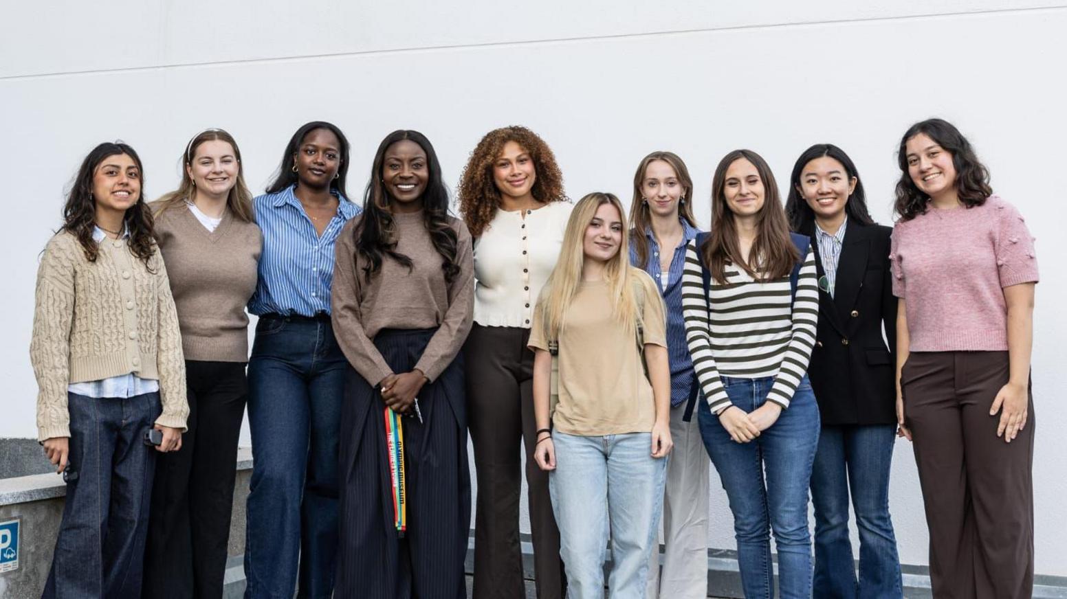 A line of 10 young women chosen for the programme, posing for a picture against a large white exterior wall. They are all smiling at the camera and have their arms down by their sides.