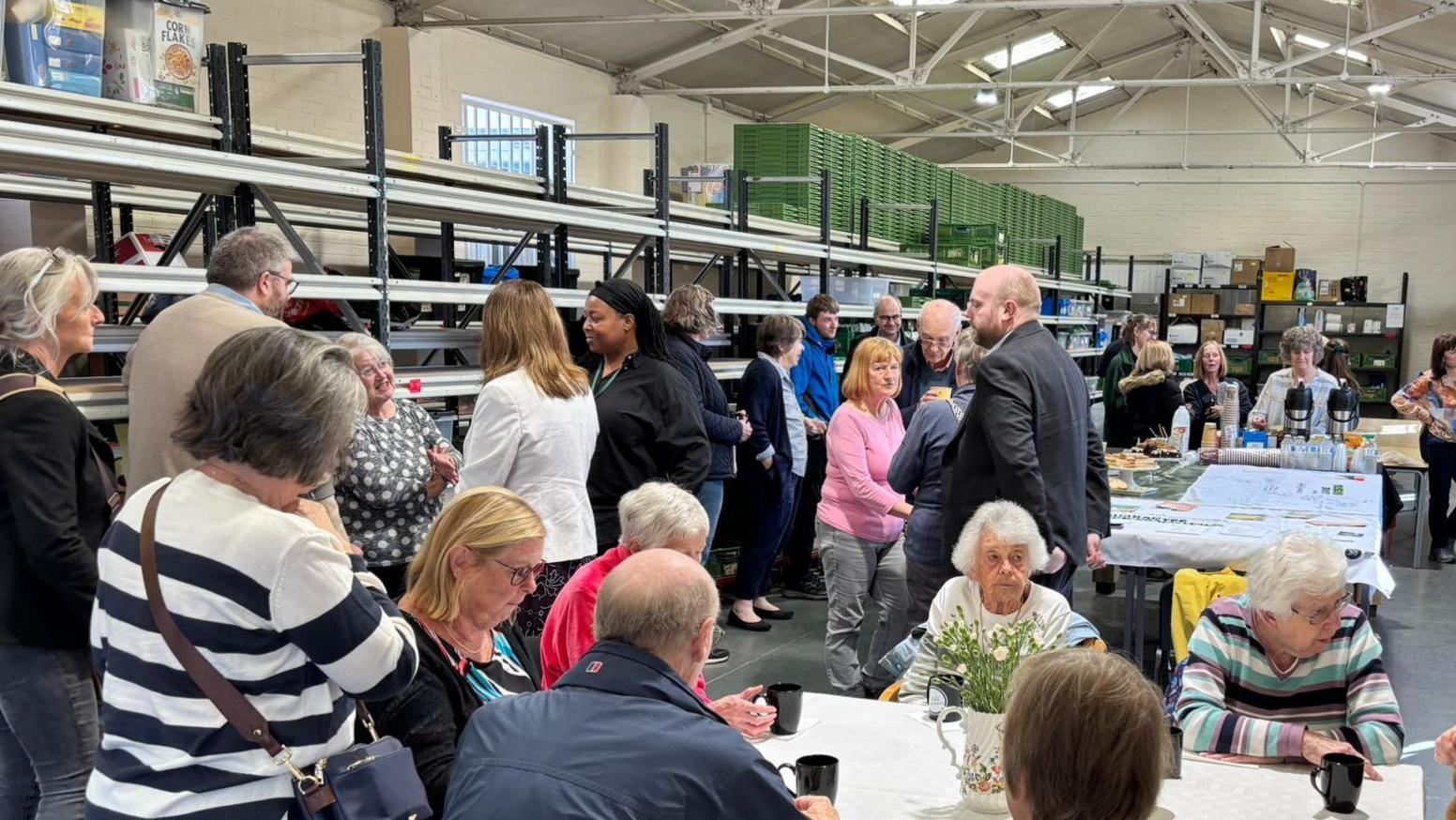 The new food bank warehouse with people milling around and sitting around tables. On the left are tall shelving units, some of which are stacked with food or toiletries items. The tables have vases with flowers and people drink cups of tea and coffee. 