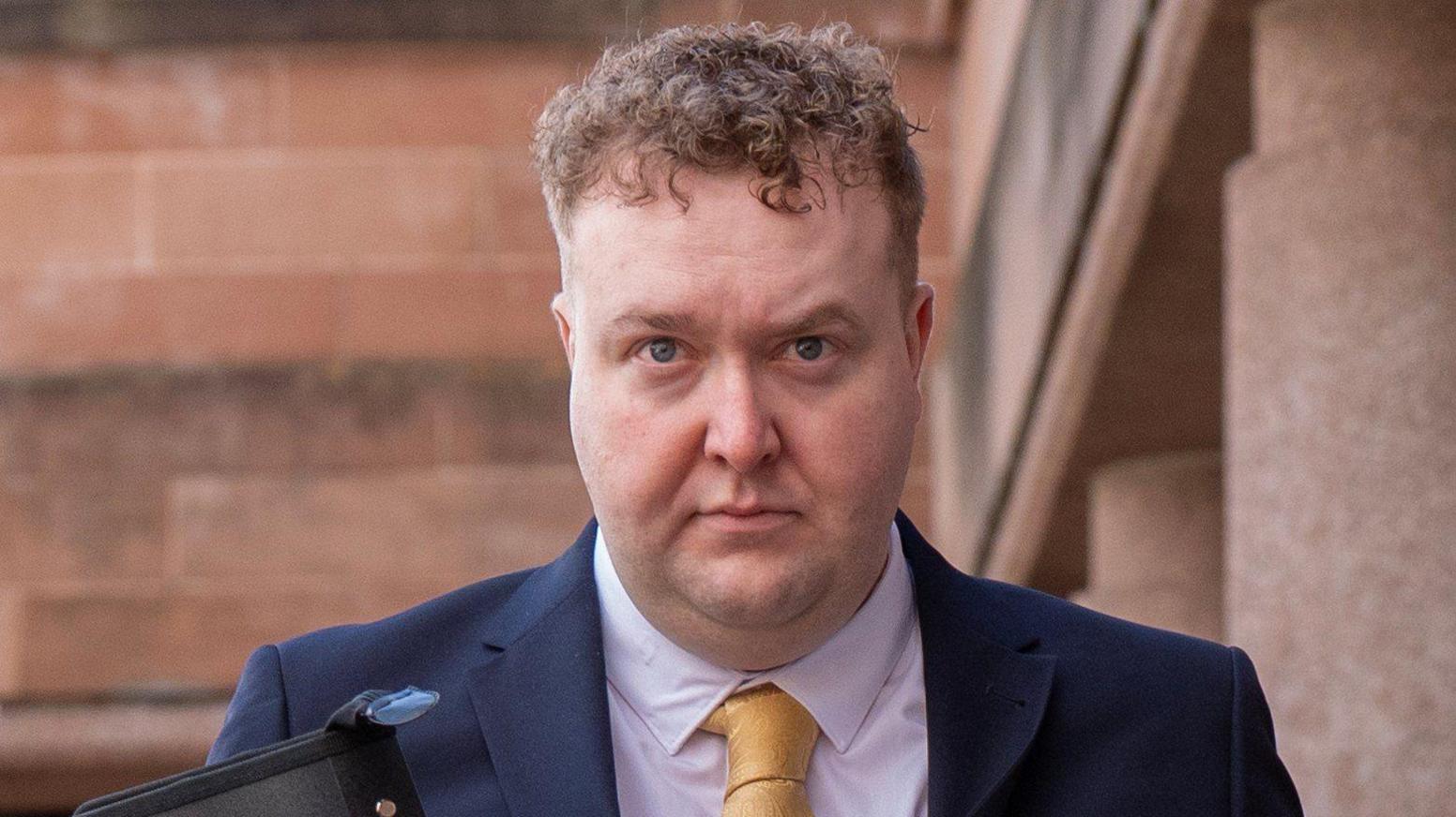A man with curly reddish brown hair, wearing a blue suit, white shirt and yellow tie looks at the camera as he leaves a court building