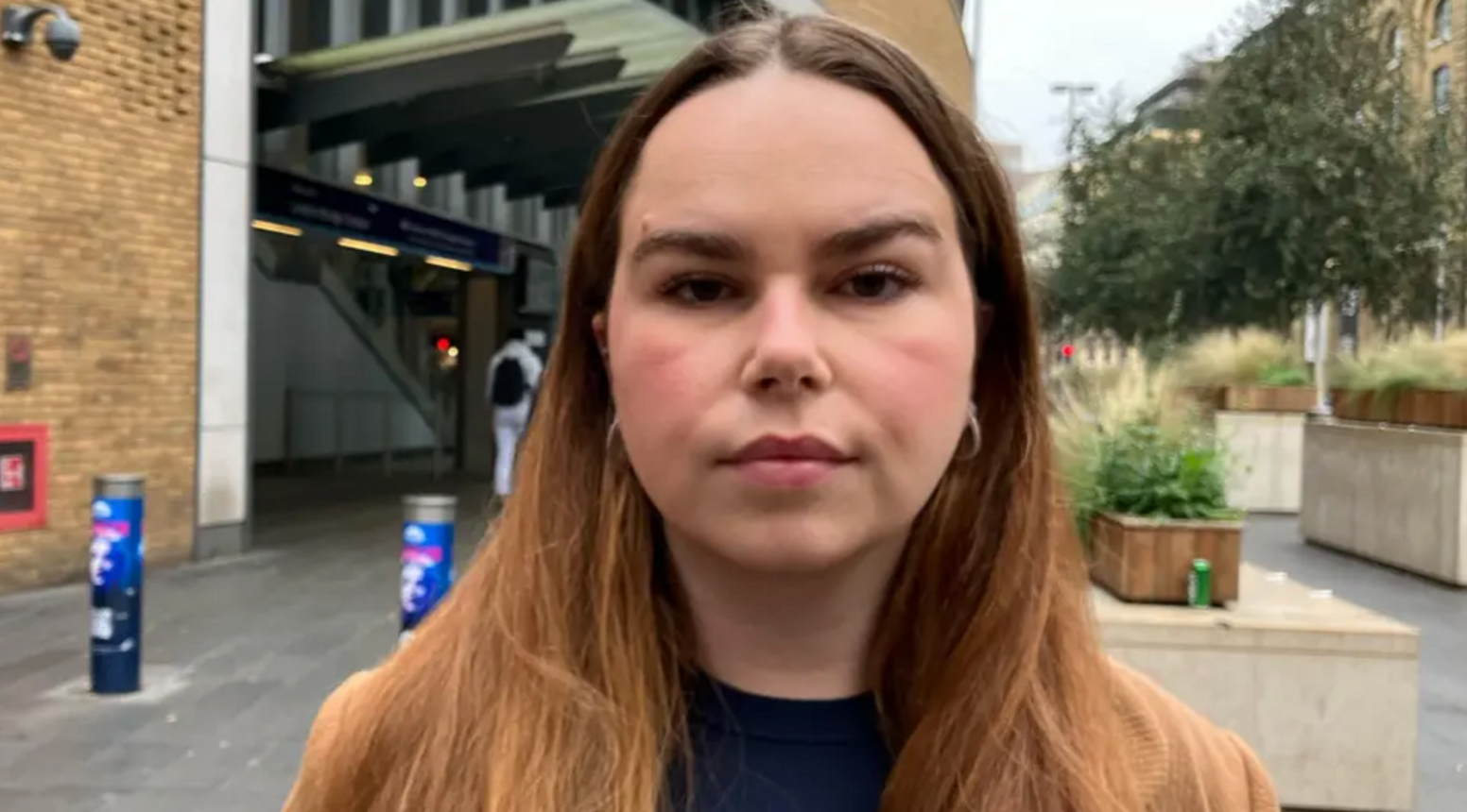 Molly looking straight at the camera. She has long straight mid-brown hair. In the background there is the entrance to a building with two large bollards  a few feet apart and some planters on the right-hand side resting on blocks of concrete.