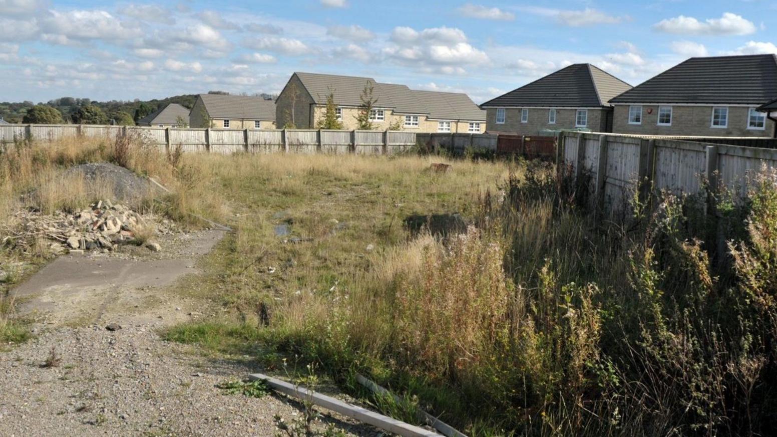 Empty grassed wasteland. Modern beige brick houses line the background behind a perimeter wooden fence.