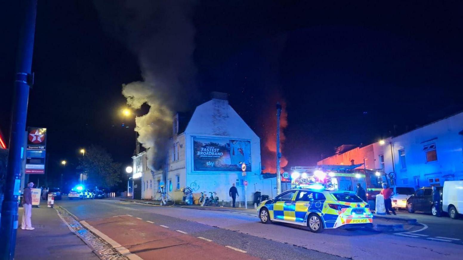 A police car is seen on a road in front of a building with smoke coming from the house. It is night time and the street lights are on.