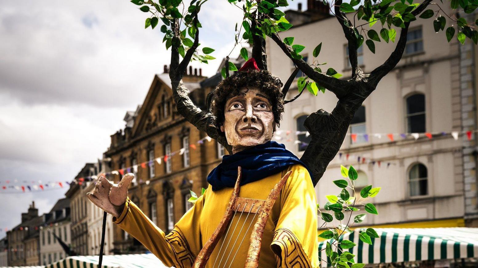 The Orpheous puppet looks towards the camera. There is papier mache foliage behind him, and he is pictured on an old city street in Cirencester lined with bunting and market stalls.
