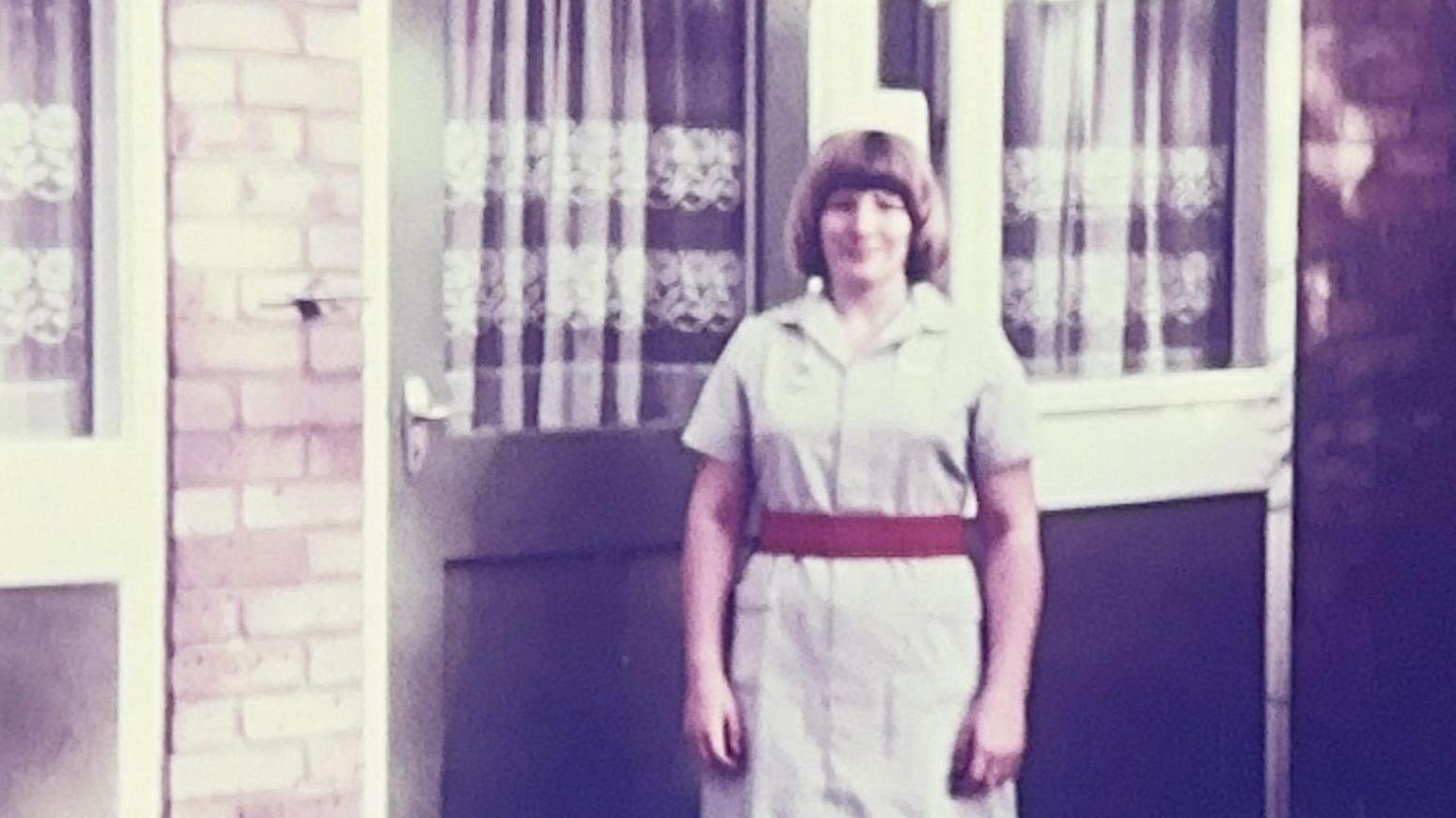 A colour photo from the 1970s of a woman standing in a white or very pale grey nurses' uniform, with a thick red elastic belt around her waist and a white box-like hat perched on top of her head. Her blonde hair is cut in a bob. She is smiling, and is outside, next to the pale green door of a house, which has a white net curtain hanging on the inside.