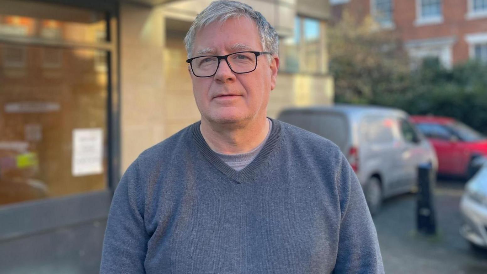 A grey haired man with black glasses stands outside wearing a blue v neck jumper.