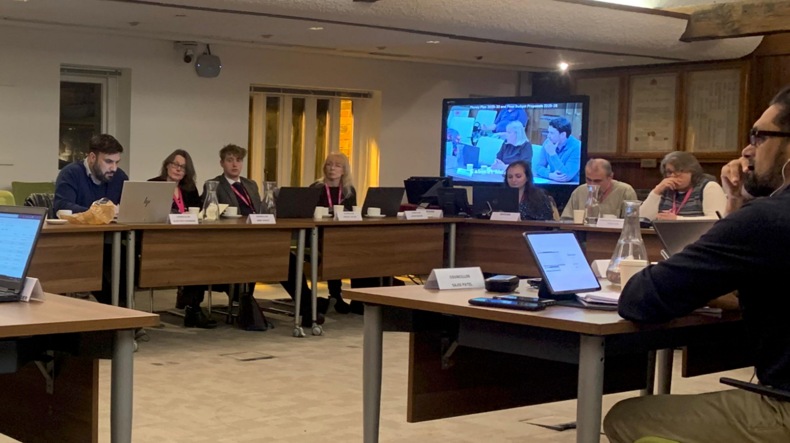 Gloucester City Councillors in a North Warehouse meeting on 20 January. There are brown desks arranged in a square format so people can face eachother. There is a large television screen in the corner of the room.