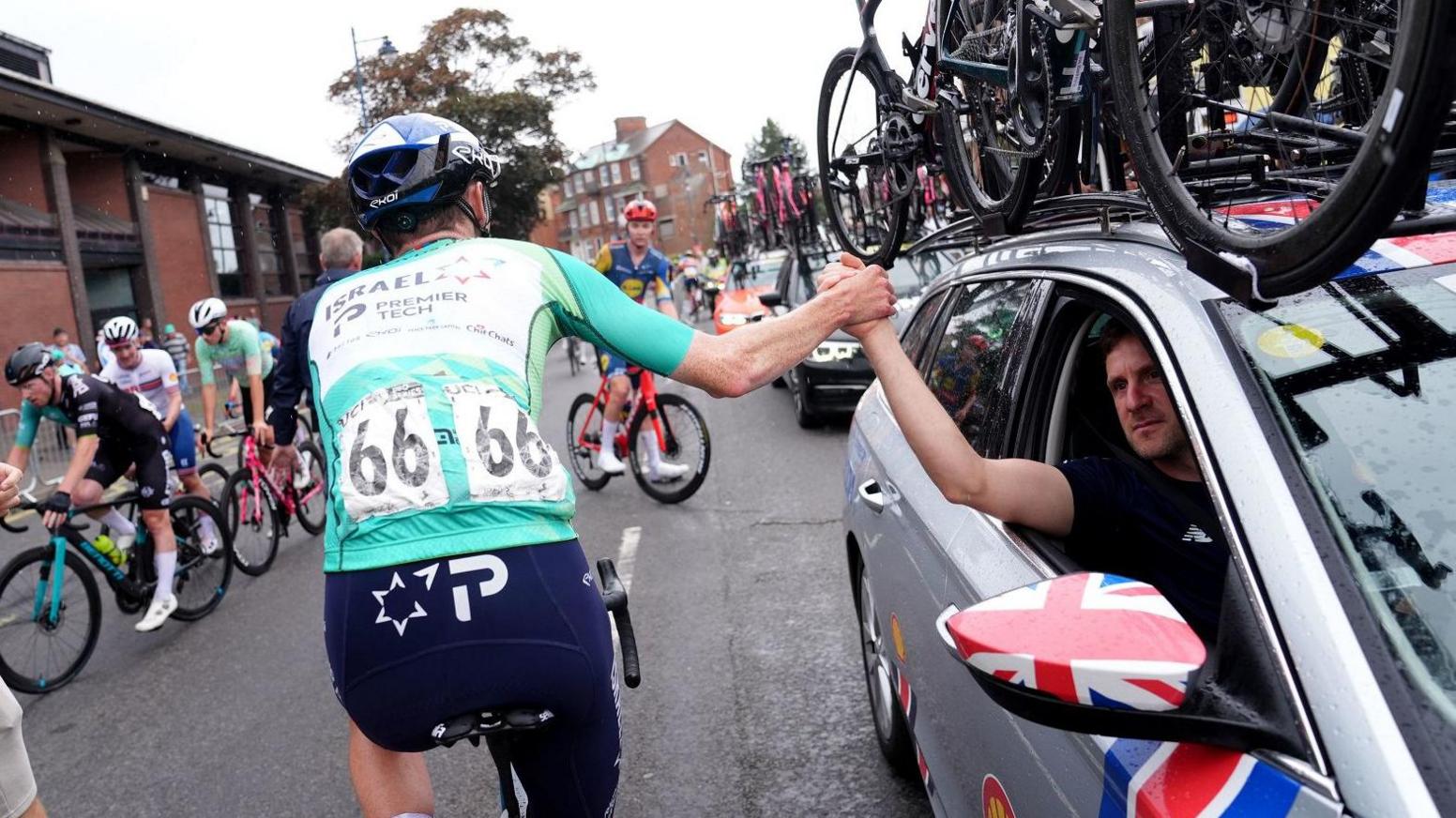 Cyclist seen from behind wearing racing tags high fiving a man driving a silver car with bikes on its roof rack, and other cyclists in the background