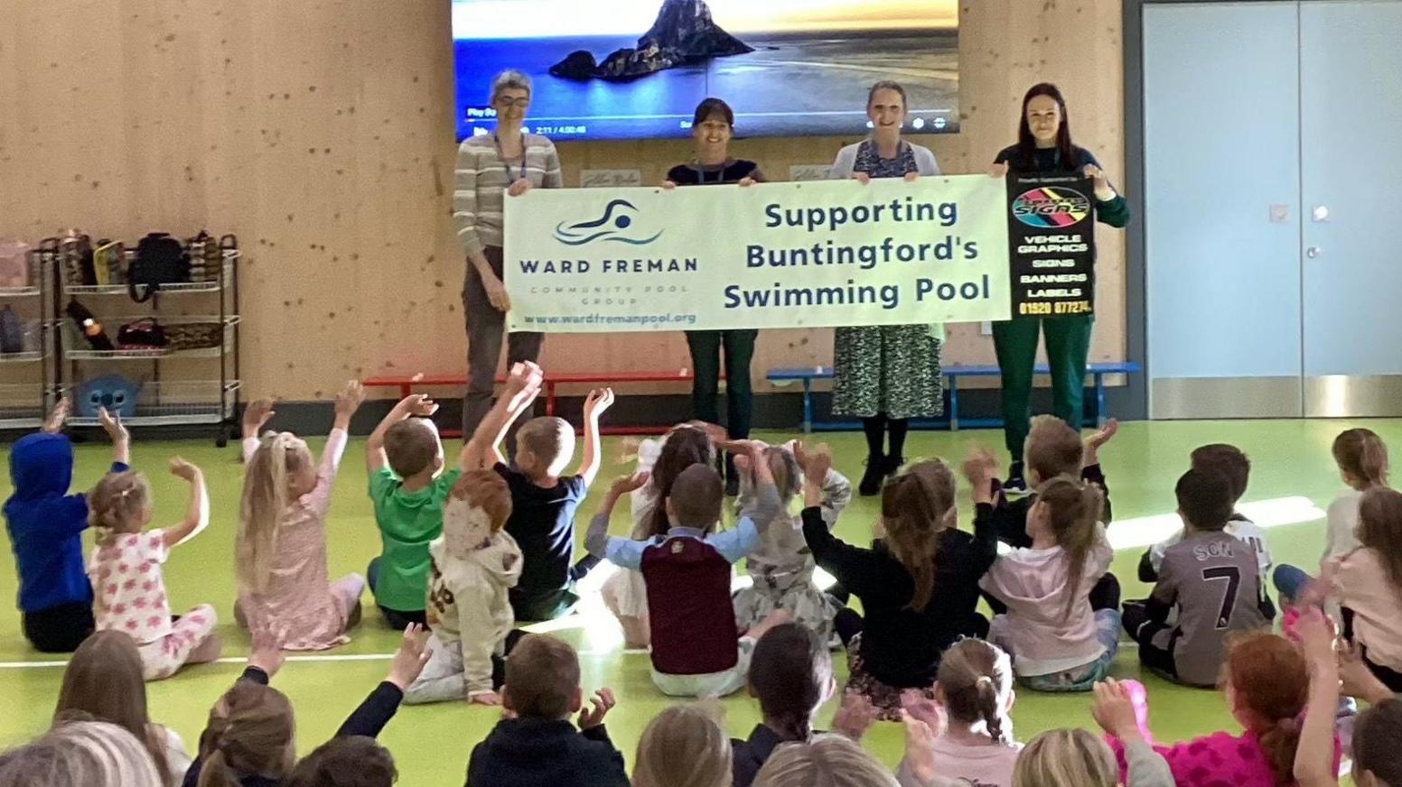 A group of school children, sitting on the floor, looking at four people who are holding a banner saying, "Supporting Buntingford's Swimming Pool"